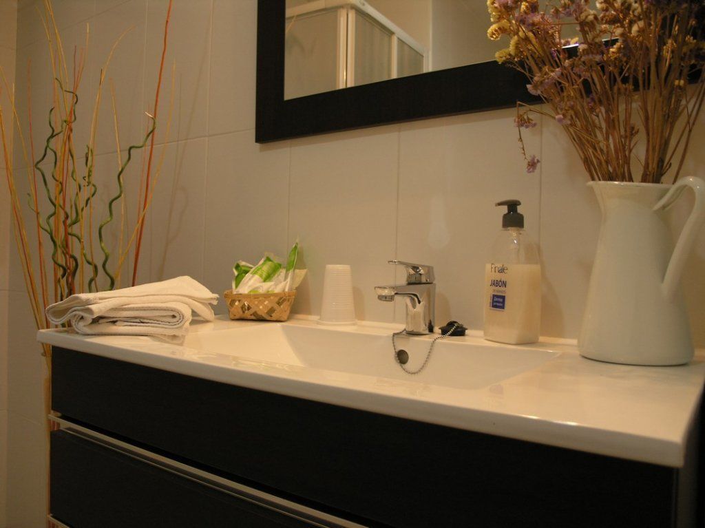 Casa de banho com lavatório branco, espelho, um frasco de sabonete e um vaso com flores secas.
