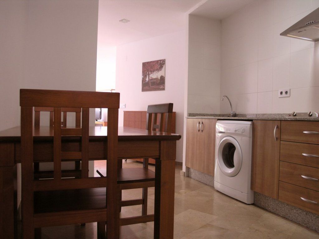 Cocina compacta con lavadora, armarios de madera y una zona de comedor con una silla.