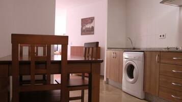 Cocina compacta con lavadora, armarios de madera y una zona de comedor con una silla.