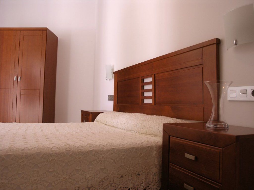 Un dormitorio con cabecera de madera, cama con cubierta de encaje, mesita de noche y armario de madera.