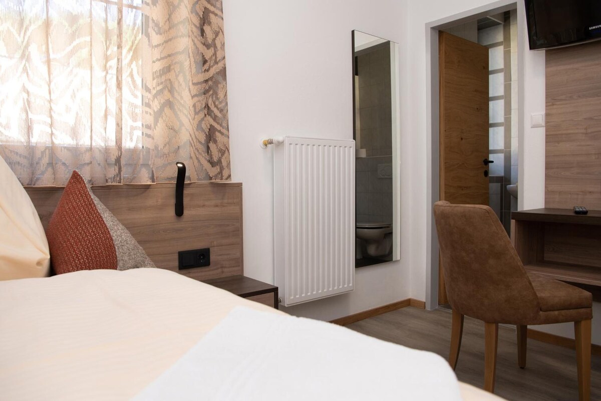 Quarto de hotel com cama, uma cadeira, radiador, espelho e televisão.