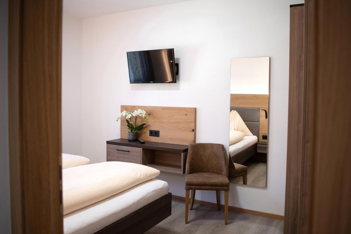 Quarto de hotel com cama, escrivaninha, cadeira, TV fixada na parede e espelho refletindo a cama e a cadeira.