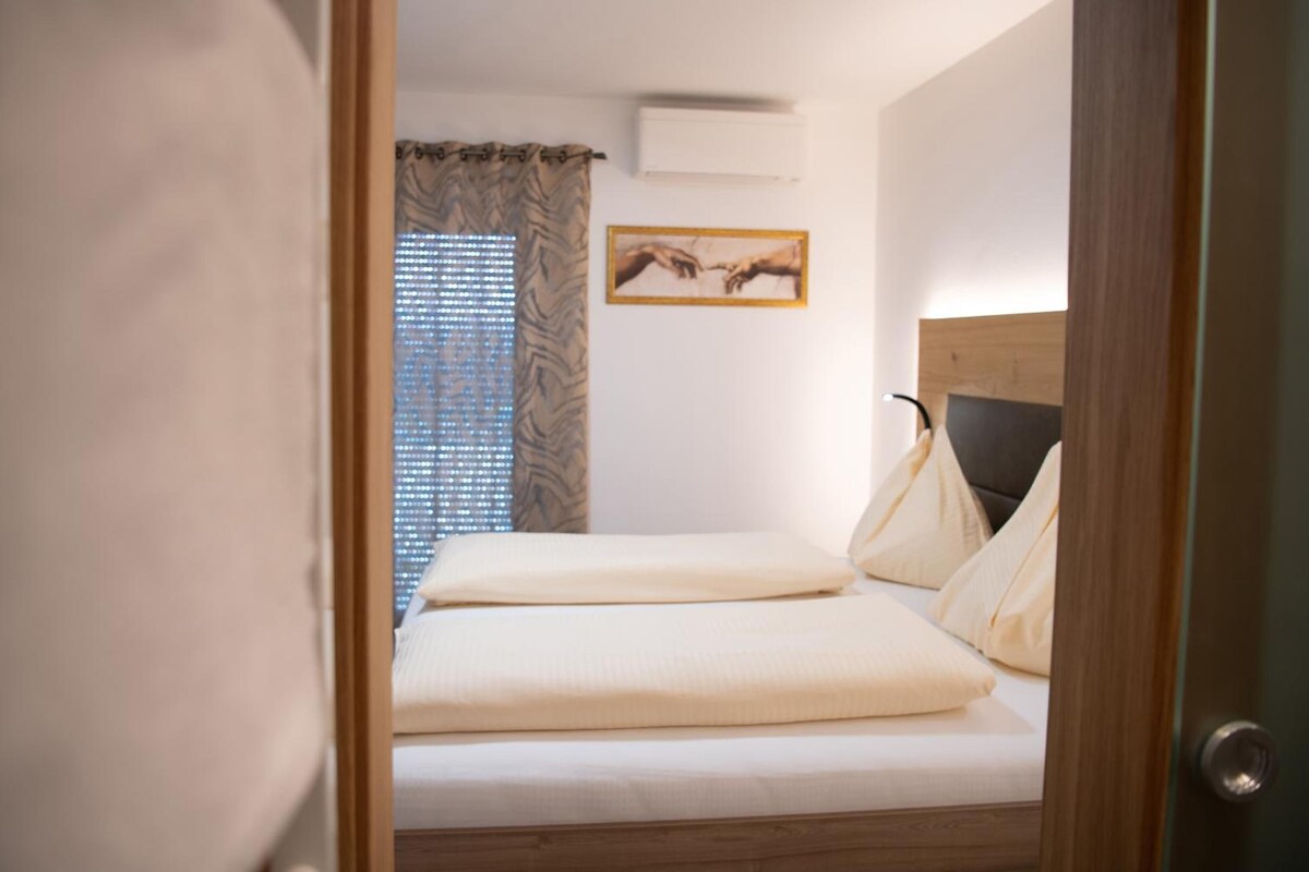 Quarto com cama, janela com persianas, um quadro na parede e uma porta.