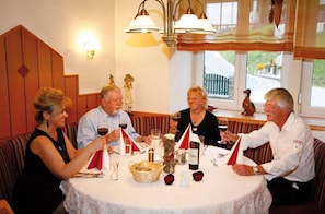 Um grupo de pessoas jantando em uma mesa redonda com toalha branca, rodeadas por velas, copos de vinho e uma cesta de pão.