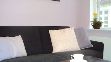 A dark grey sofa with patterned cushions, a white cup and saucer on a wooden table, and a window with a view of buildings and a plant on the windowsill.