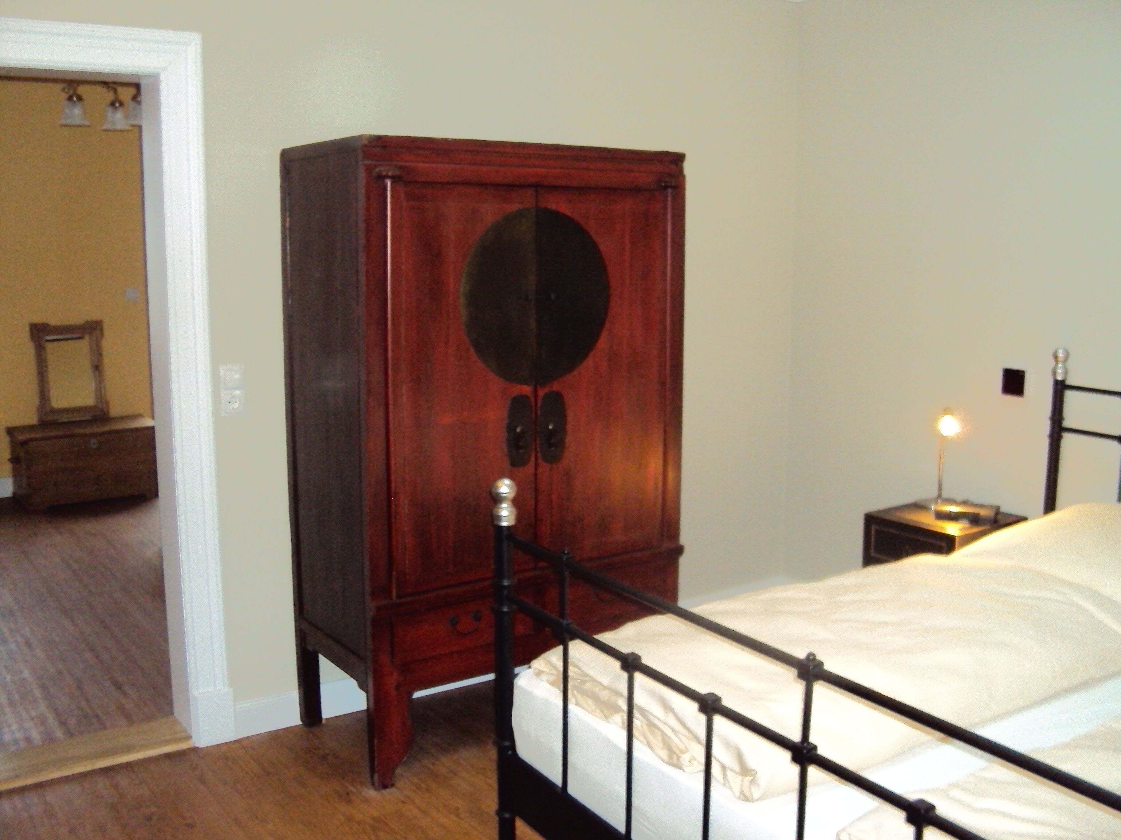 A bedroom with a wooden wardrobe, a bed with a metal frame, a nightstand with a lamp, and a mirror on the wall.
