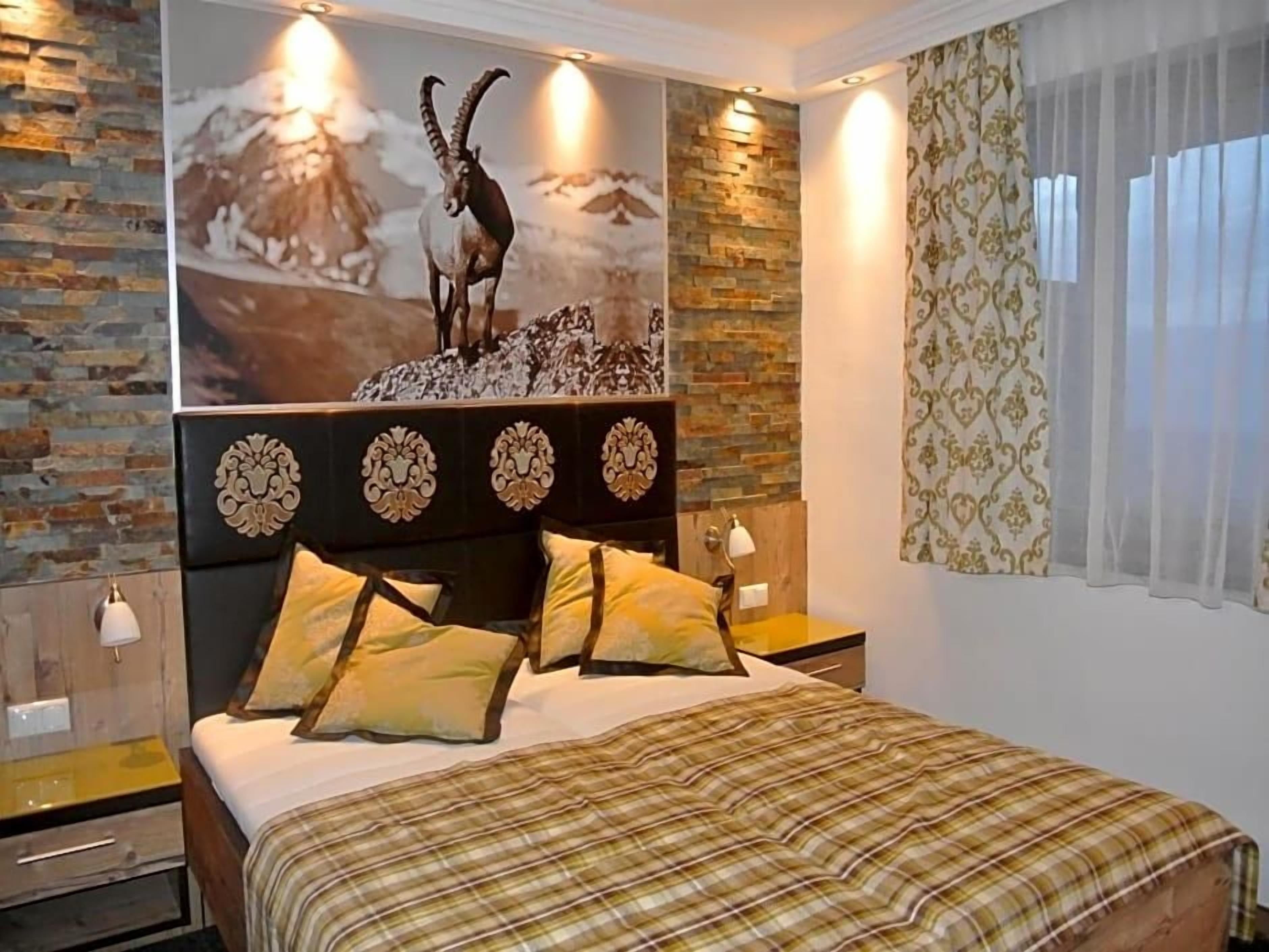 A bedroom with a bed, a headboard with decorative panels, a nightstand, a wall-mounted lamp, and a window with curtains.