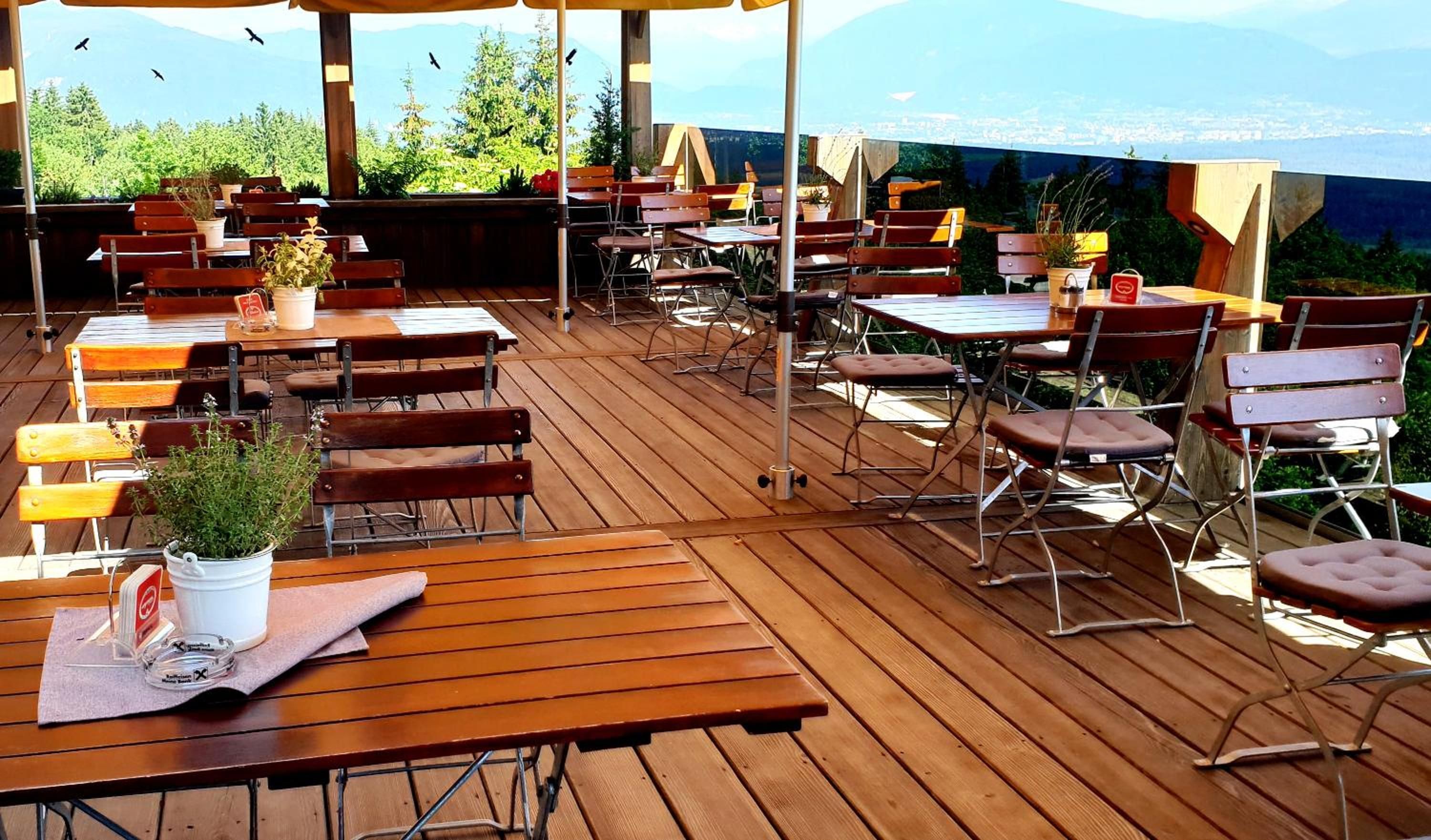 Een terras met houten tafels en stoelen, plantenbakken en uitzicht op de stad.