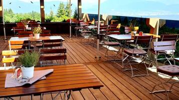 Een terras met houten tafels en stoelen, plantenbakken en uitzicht op de stad.