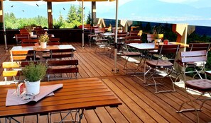 Un área de comedor al aire libre con mesas y sillas de madera, plantas en macetas y vistas de la ciudad.