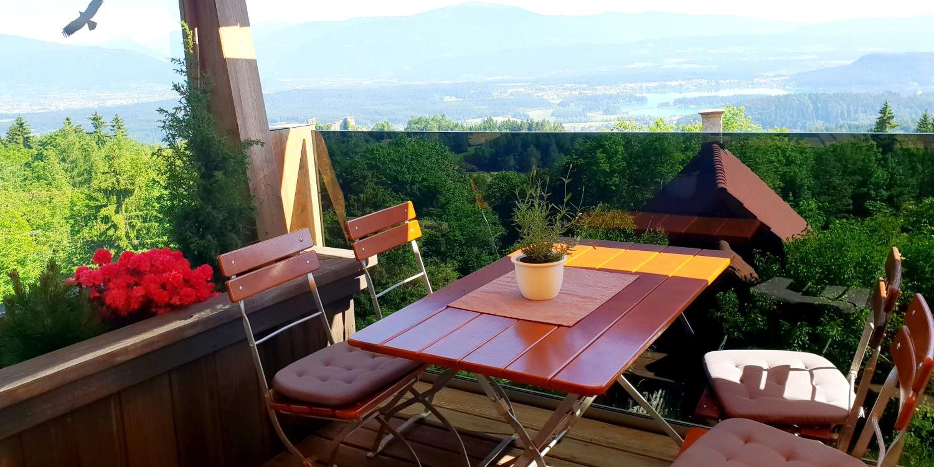 Eine Holzterrasse mit Tisch und Stühlen, einem Blumentopf und Blick auf einen Wald und ferne Berge.