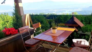 Eine Holzterrasse mit Tisch und Stühlen, einem Blumentopf und Blick auf einen Wald und ferne Berge.