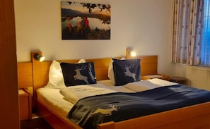 A bed with two decorative pillows, a bedside table, a nightstand, a wall-mounted lamp, and a painting above the bed. - Baumgartnerhof (Finkenstein am Faaker See)