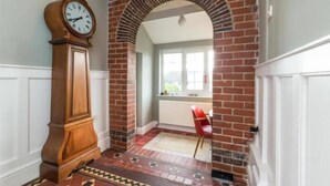 A hallway with a brick archway, a wooden floor with patterned tiles, a red chair, and a large wall clock.