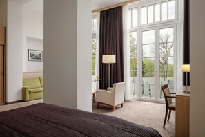 A hotel room with a large bed, a green armchair, a wooden desk, and a view of the outdoors through large windows. - Harvey SPA & KUR (Frantiskovy Lazne)