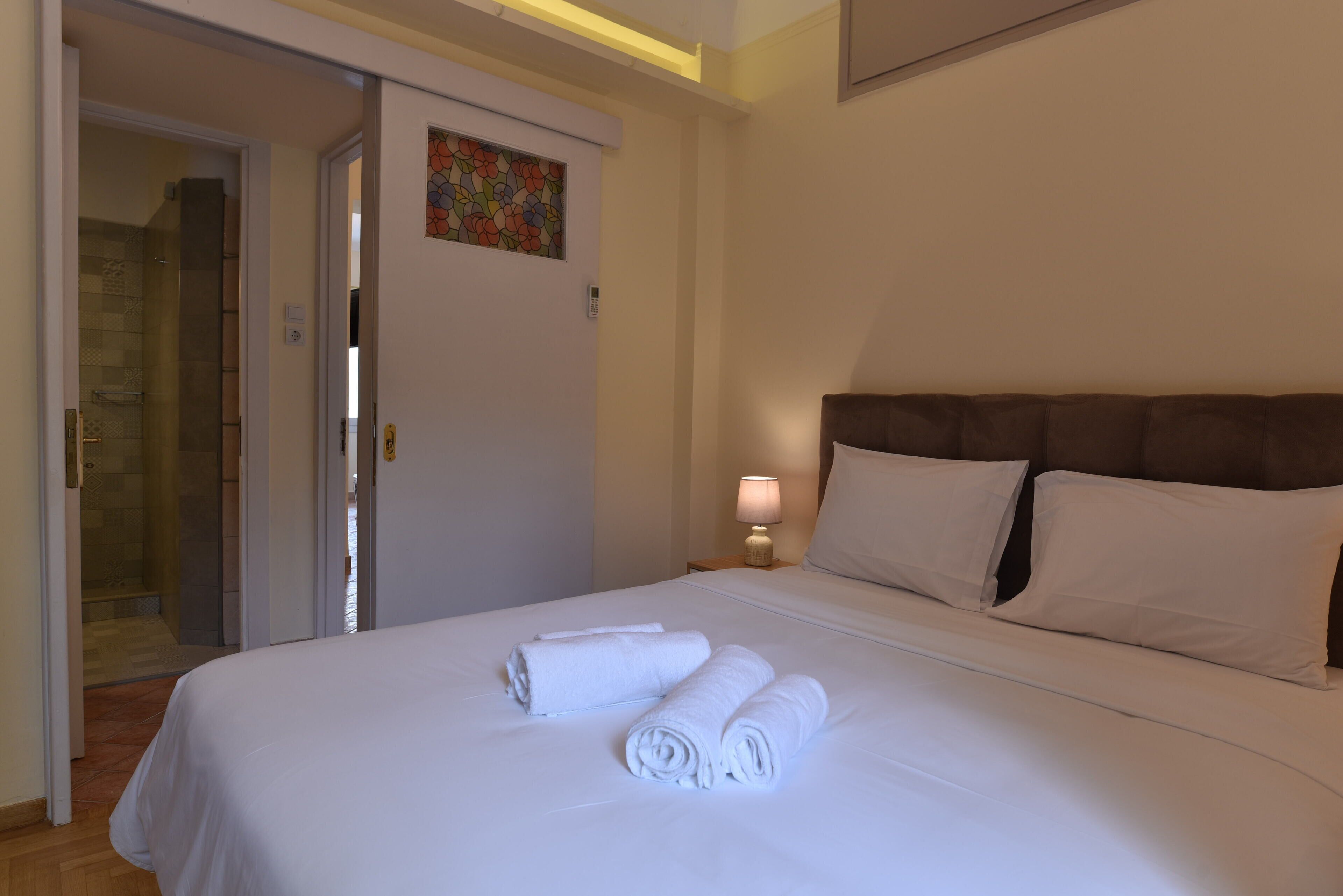 Habitación de hotel con una cama bien hecha, una puerta que da al baño y una mesita de noche pequeña con una lámpara.