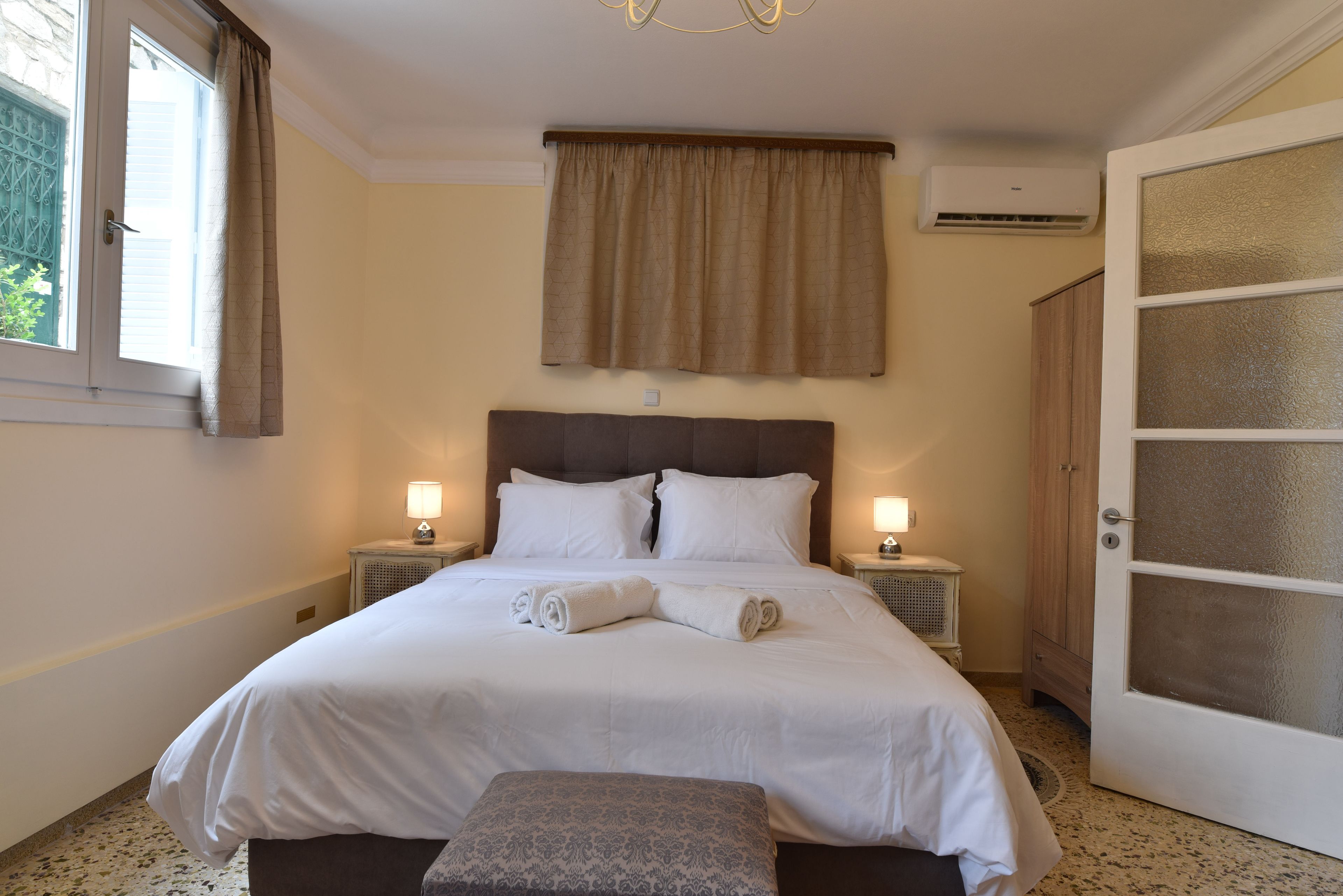 Una cama bien hecha con sábanas blancas, dos lámparas de noche, una ventana con cortinas y una puerta con paneles de vidrio esmerilado.
