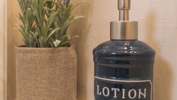 Una botella de loción con dosificador de bomba sobre una estantería, junto a una planta en maceta con flores moradas.