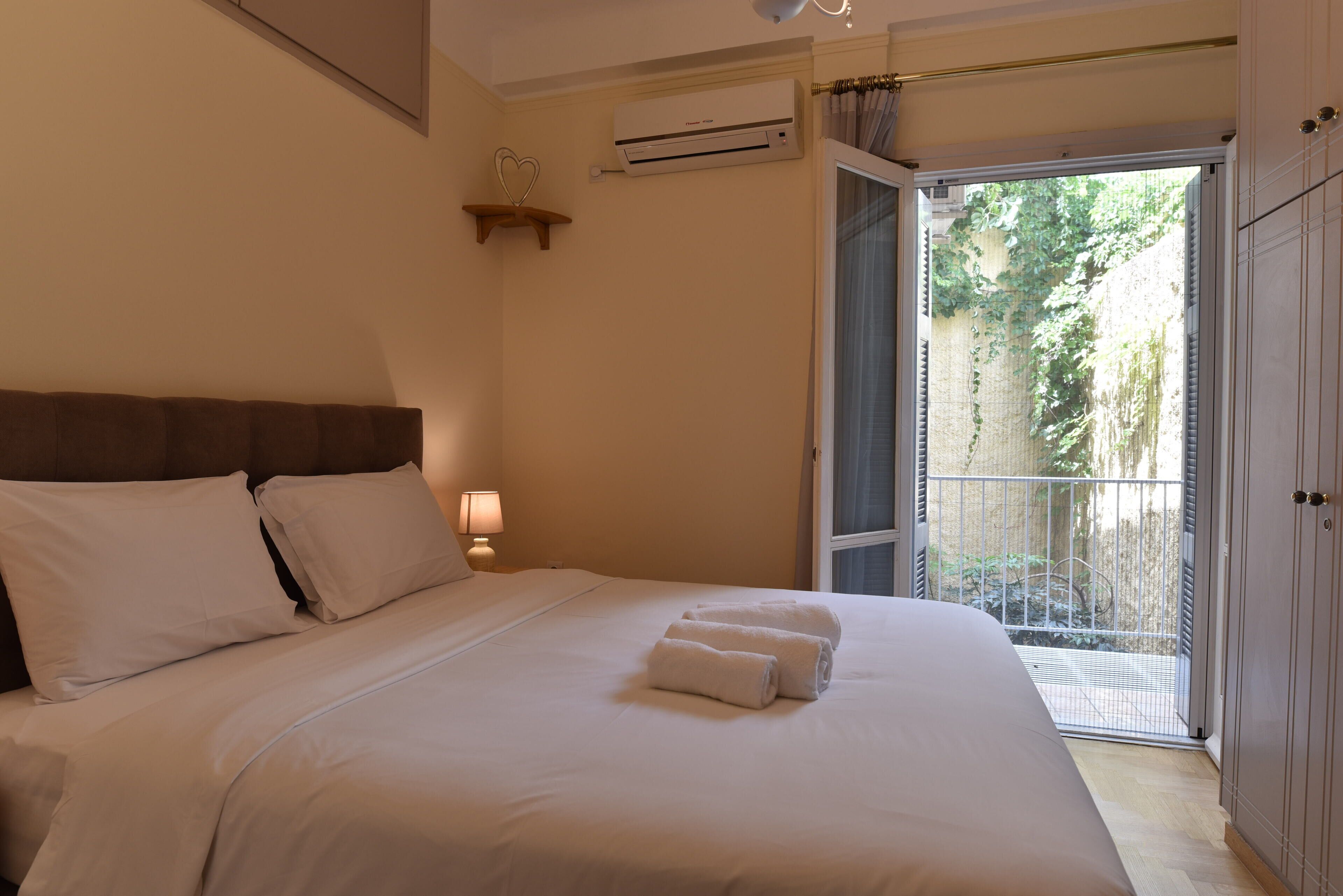 A bedroom with a bed, a nightstand, a lamp, an air conditioning unit, and a balcony with a view of greenery.