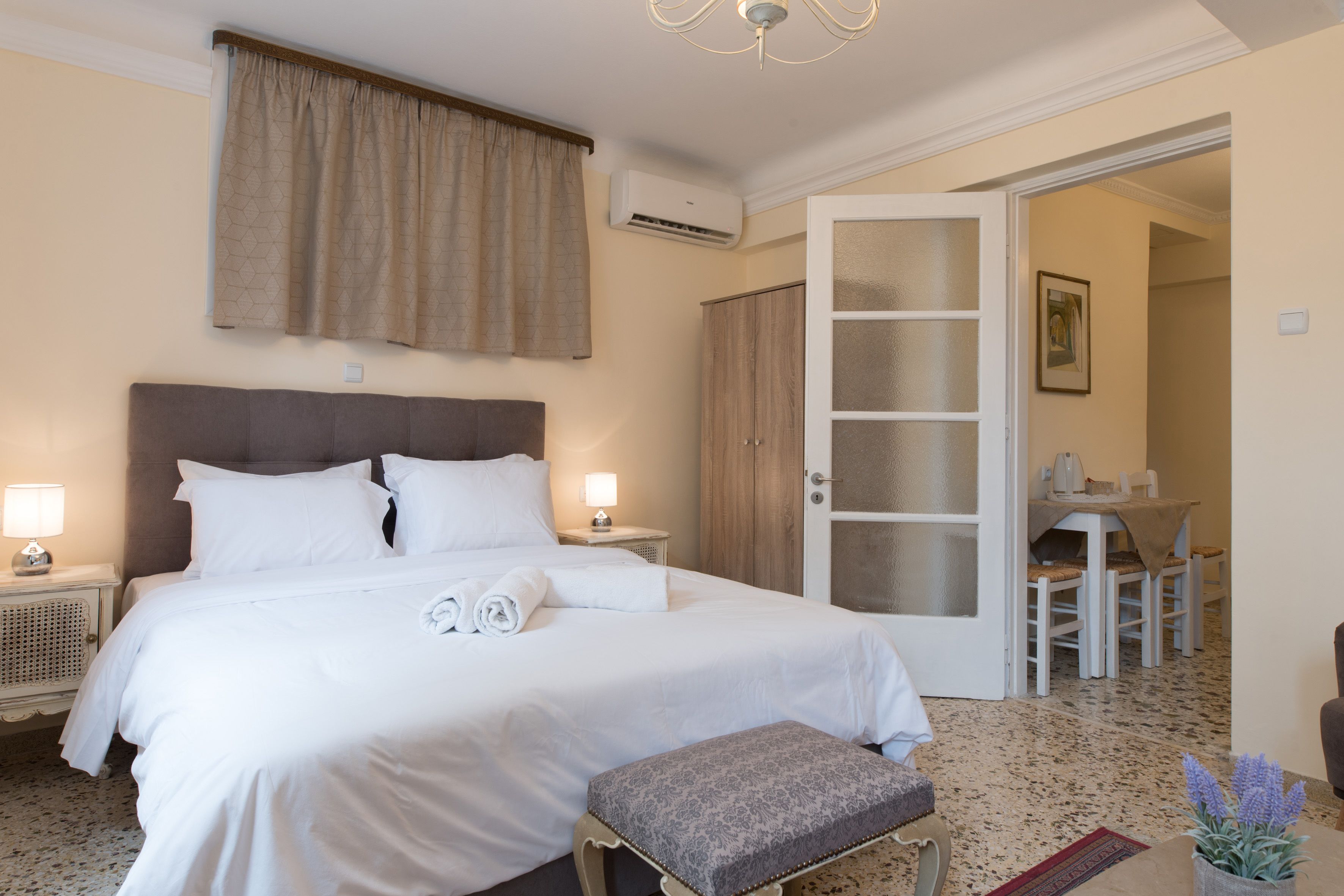 A hotel room with a large bed, bedside lamps, a small table, and a door leading to a dining area.