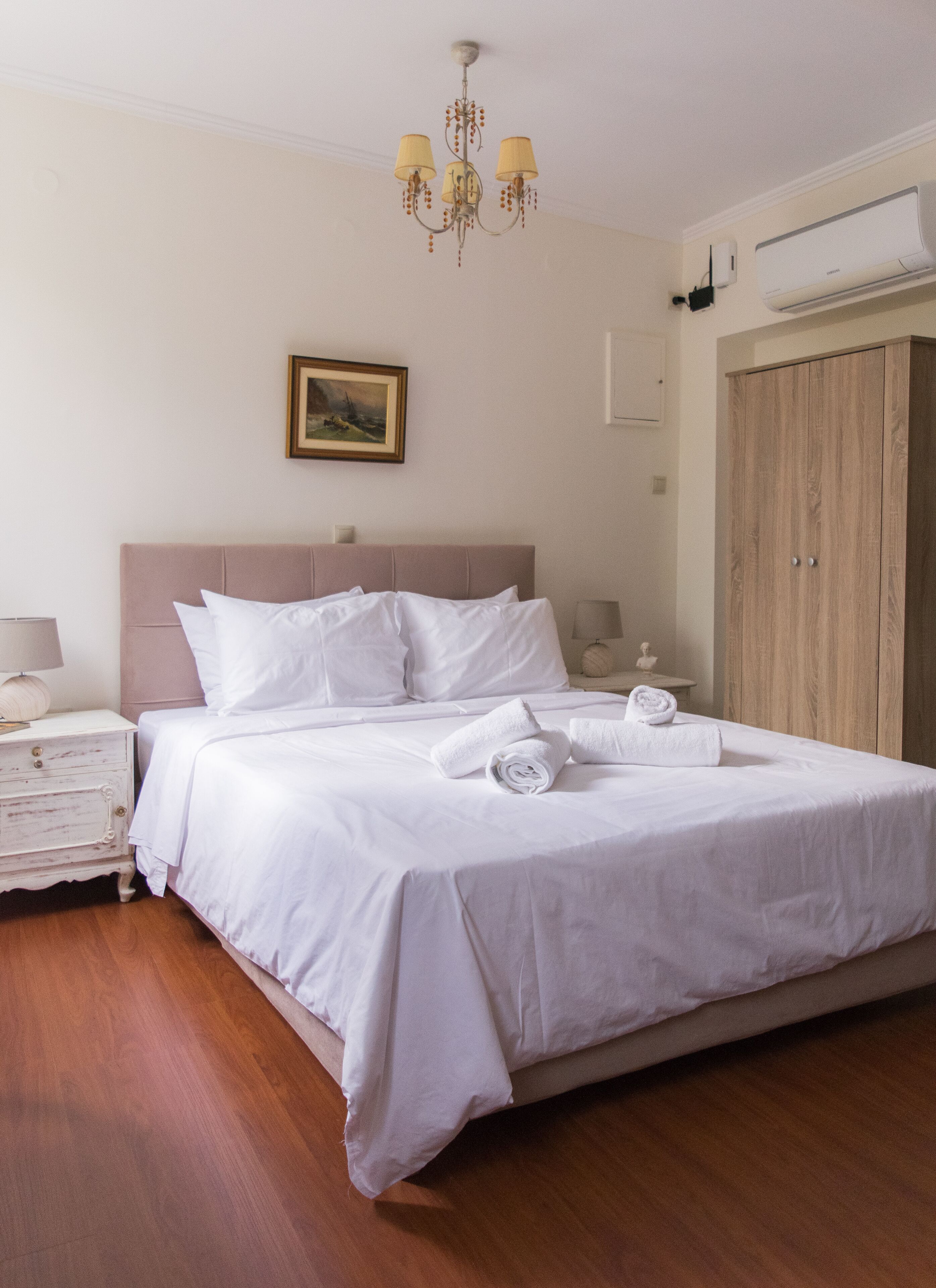 A neatly made bed with white linens, a wooden headboard, a bedside table with a lamp, and a painting on the wall.