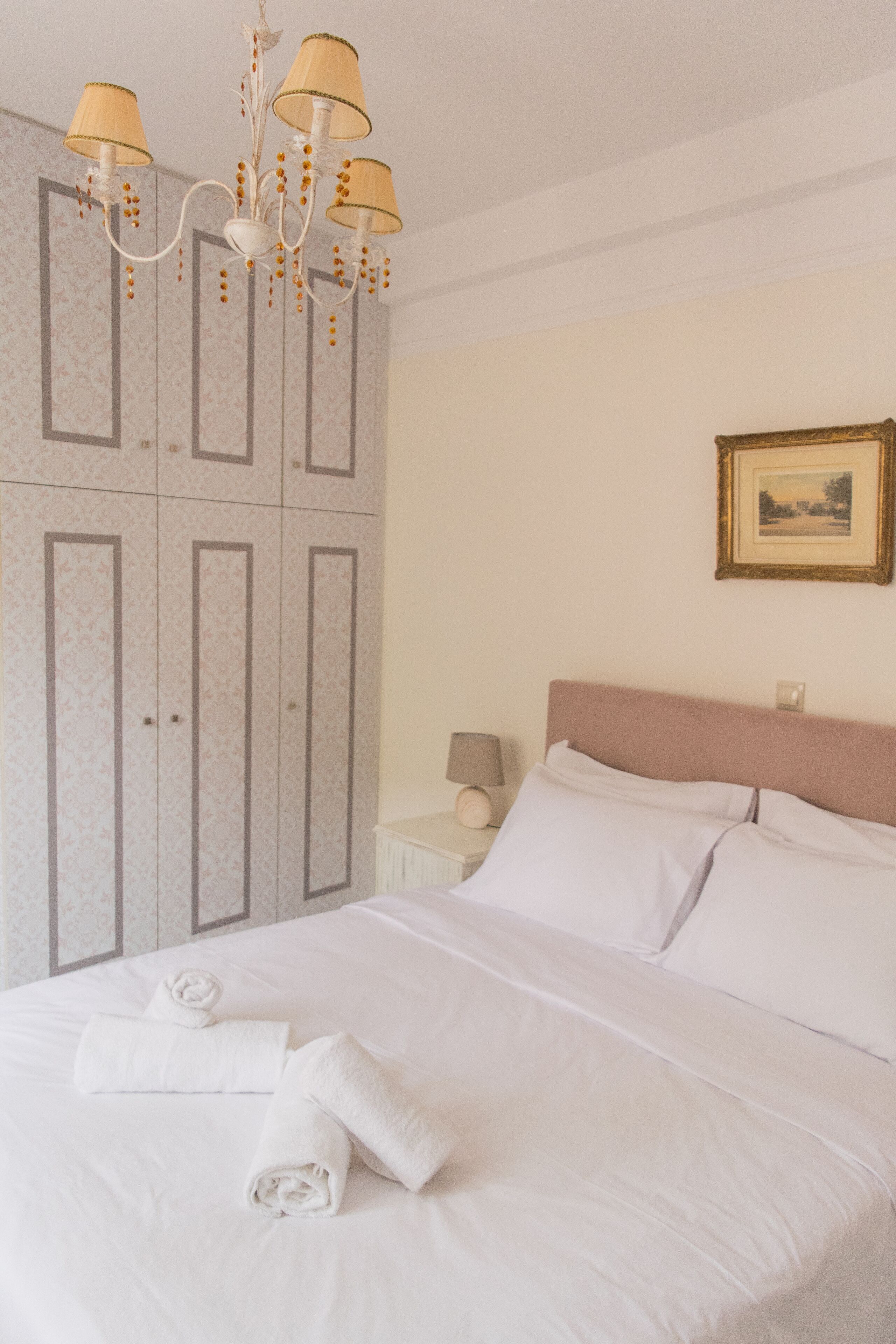 A neatly made bed with white linens and rolled towels, a chandelier with beige lampshades, a nightstand with a lamp, and a framed picture on the wall.