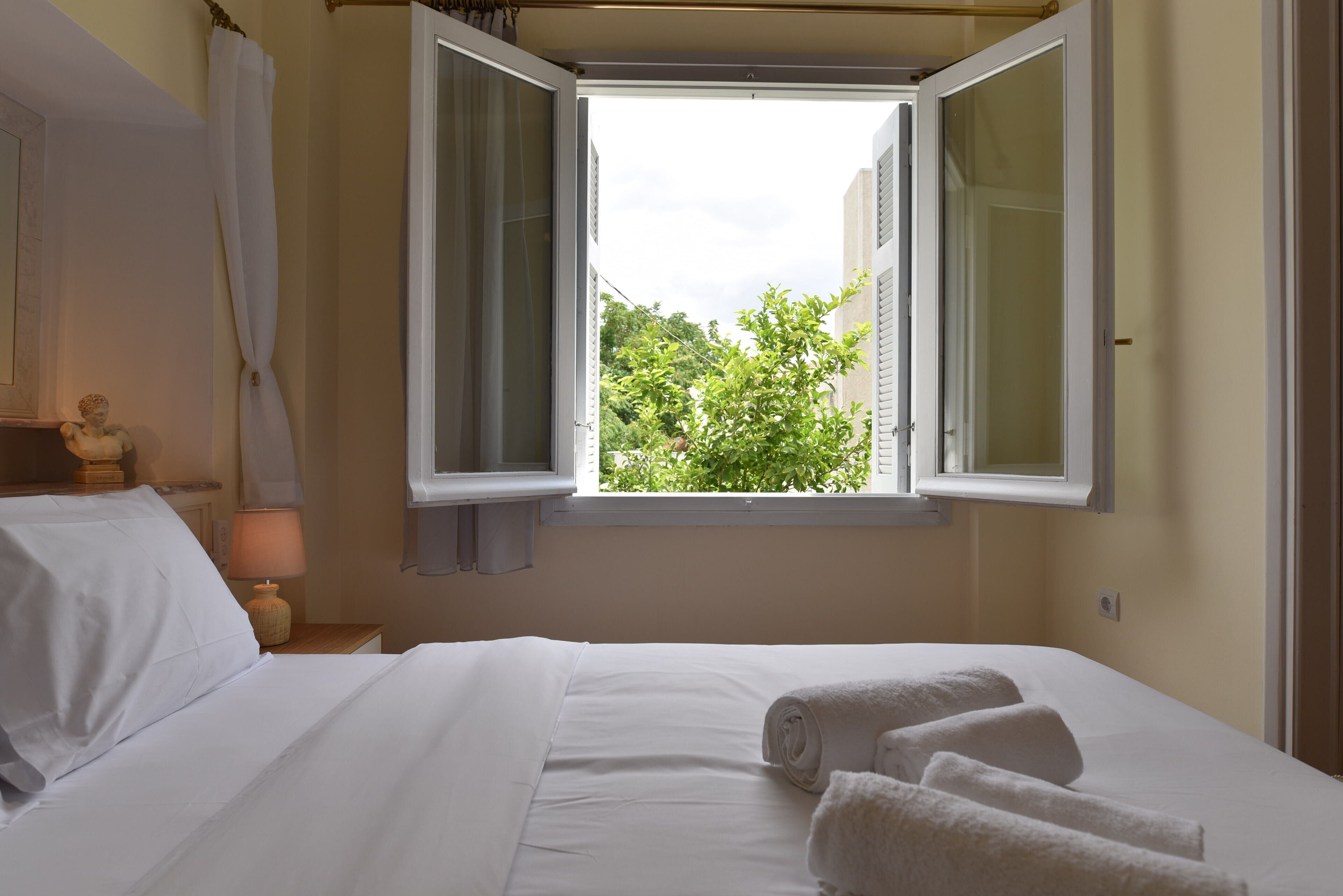 Une chambre à coucher avec un lit, du linge de lit blanc, une fenêtre avec des volets ouverts, une table de chevet avec une lampe et une vue sur la verdure à l’extérieur.