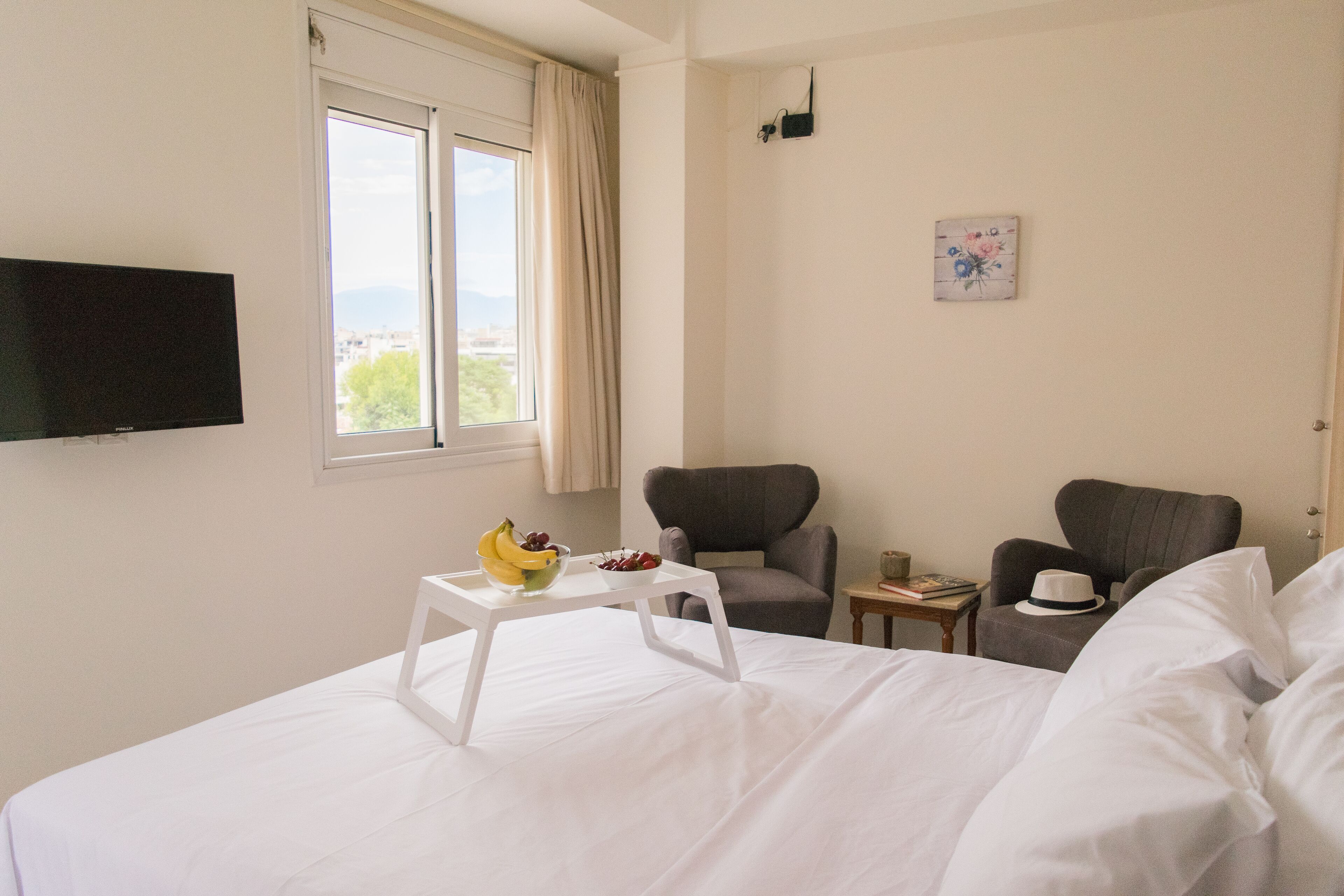 Una habitación de hotel con una cama, dos sillones, una mesita con un plátano y un cuenco de fruta, un televisor montado en la pared y una ventana con cortinas.