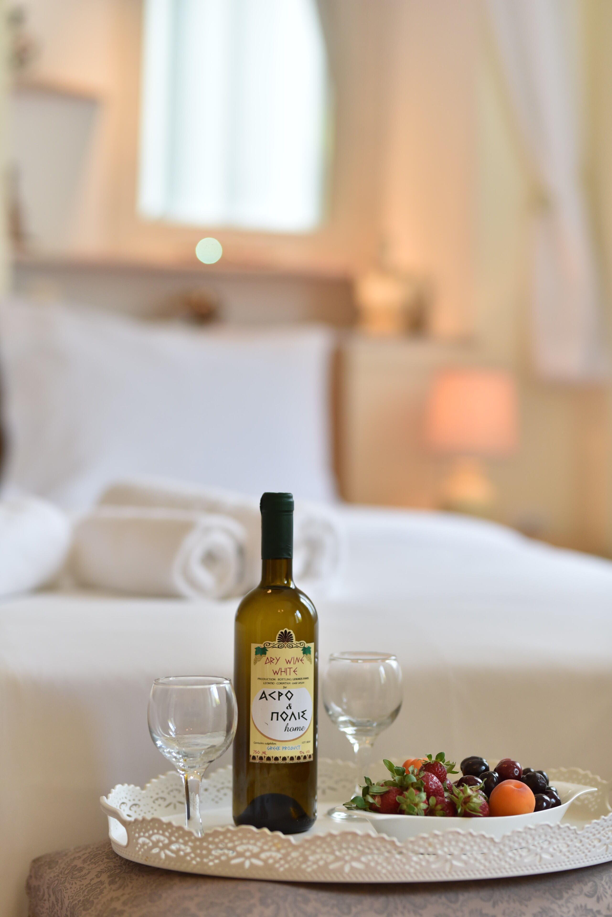 Un lit bien fait, avec une bouteille de vin, deux verres et une assiette de fruits sur une table.