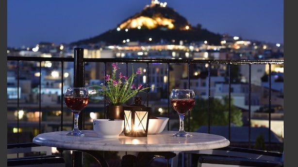 Une table avec deux verres à vin et une plante en pot, sur un balcon donnant sur une vue nocturne de la ville.