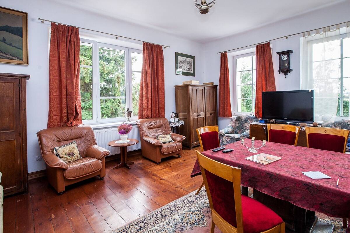 Ein gemütliches Wohnzimmer mit Holzboden, Ledersesseln, einem Esstisch mit roter Polsterung und einem Fernseher.