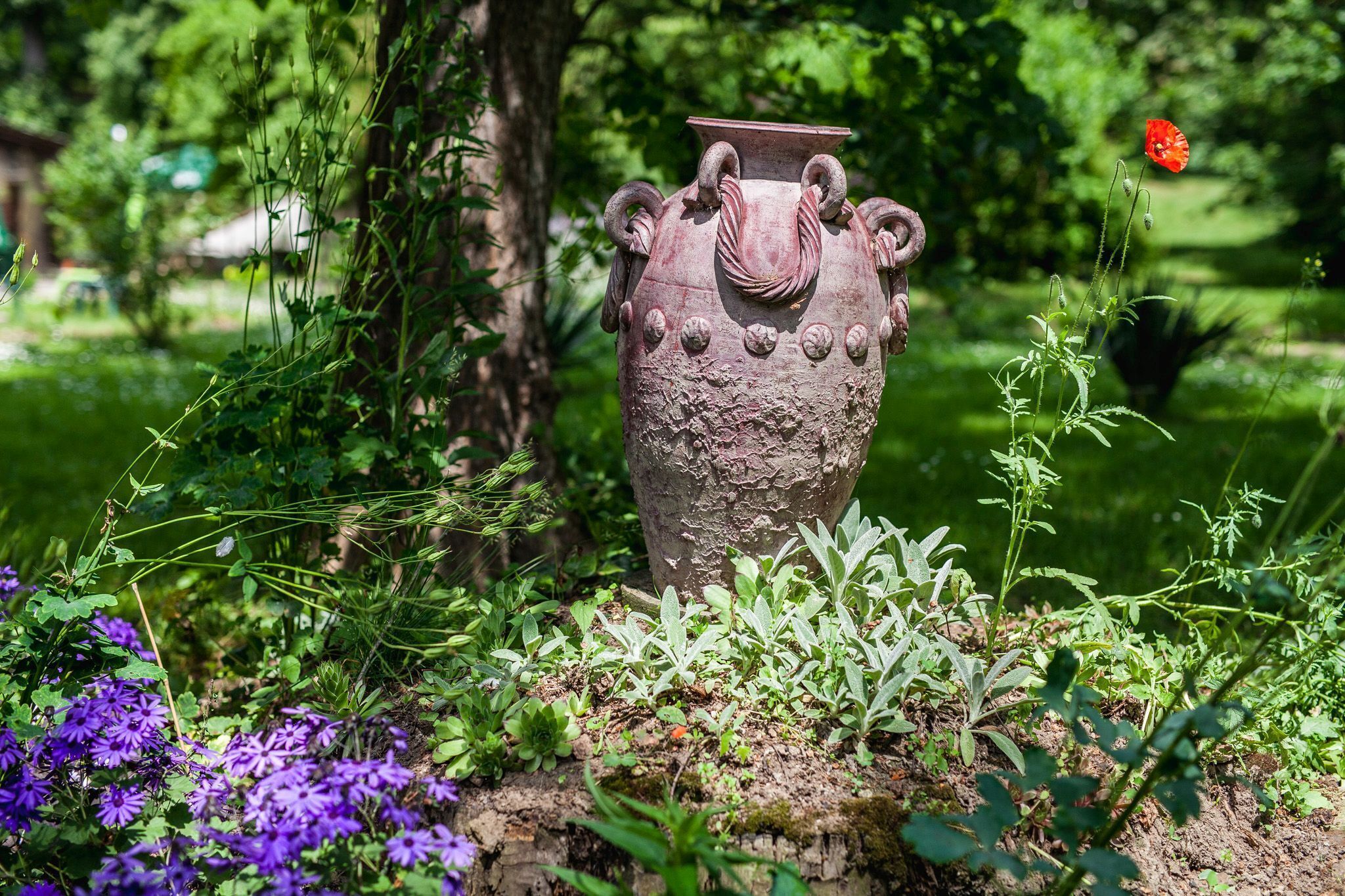 Een rustieke stenen vaas in een tuin, omringd door groen en een boom.