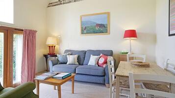 Uma sala de estar acolhedora com um sofá cinzento, uma mesa de centro de madeira, uma mesa de jantar com cadeiras brancas, um abajur vermelho e um quadro com vacas na parede.