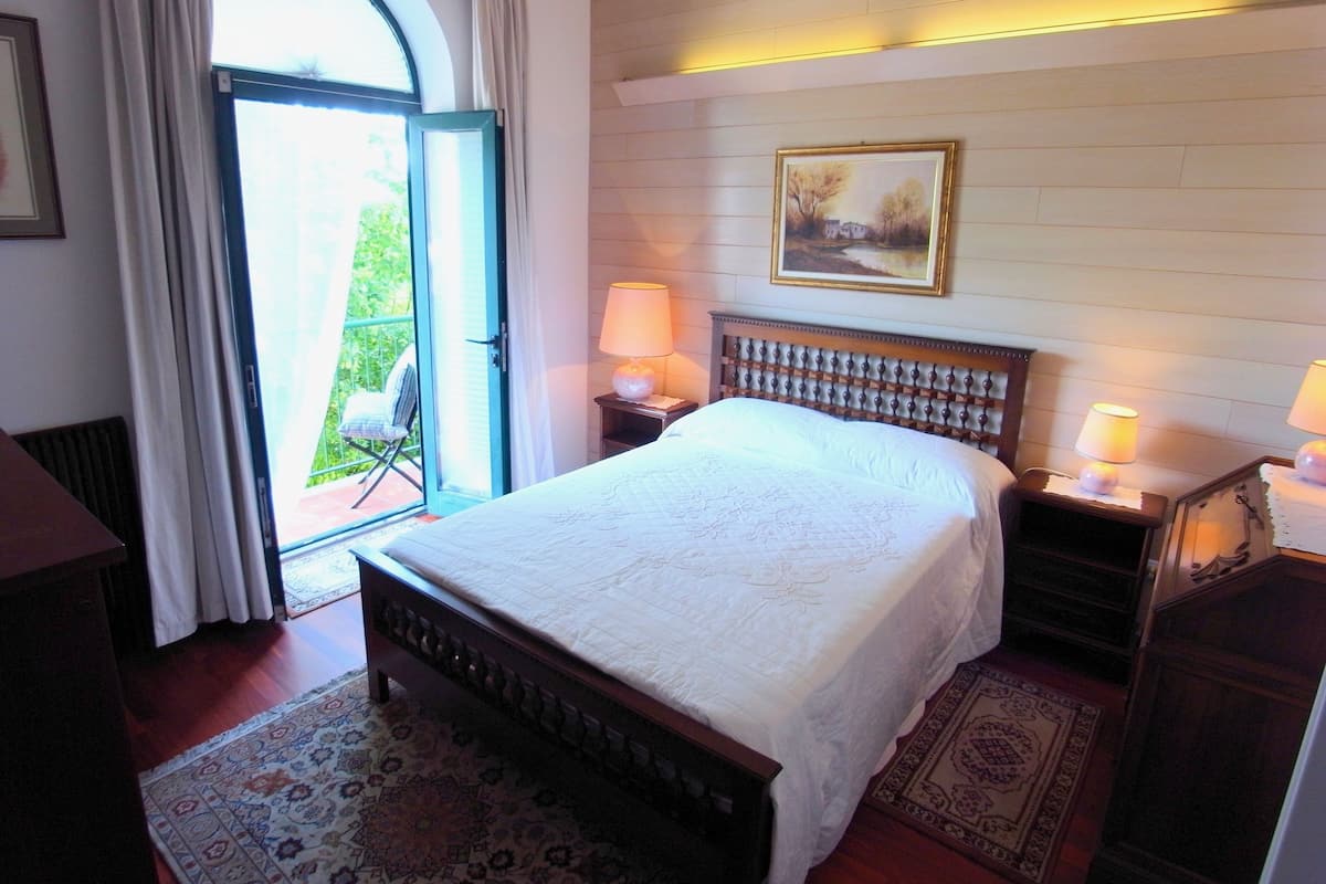 A bedroom with a large bed, two bedside tables with lamps, a painting on the wall, and a view through a glass door.
