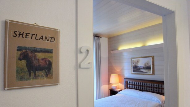 Une chambre d’hôtel avec un lit, une table de chevet avec une lampe, un tableau encadré au mur et une enseigne portant le nom « Shetland ».