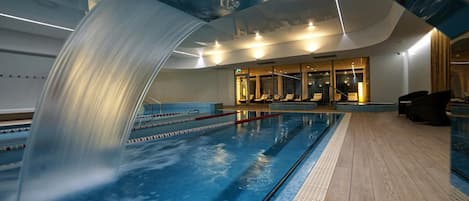 Une piscine agrémentée d’une cascade, un espace détente avec des sièges et un mur entièrement vitré.