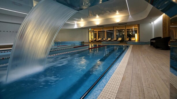 A swimming pool with a waterfall feature, a lounge area with seating, and a glass-enclosed wall. - Hotel Ba Antowo Sport (Katowice)