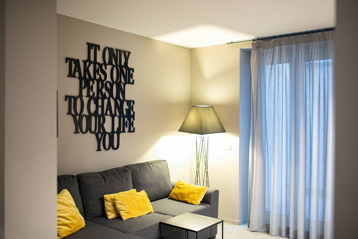 A modern living room with a grey sofa, yellow cushions, a floor lamp, and a large motivational quote on the wall.