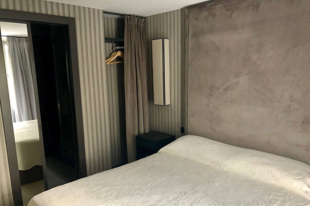 A hotel room with a bed, a bedside table, a hanging coat rack, a mirror, and a window with curtains.