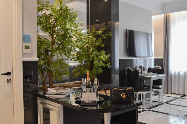Una habitación de hotel moderna con encimera de mármol negro, un gran árbol en maceta, una zona de comedor con mesa y sillas, y un televisor de pantalla plana montado en la pared.