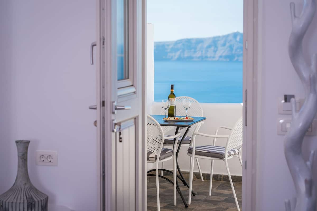 An open door leading to a balcony with a table set for two, a bottle of wine, and a view of the sea and cliffs.