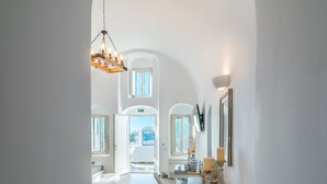 A hallway with two large vases on a white console table, leading to a bright room with a view of a blue gate. - Angel Luxury Suites (Santorini)