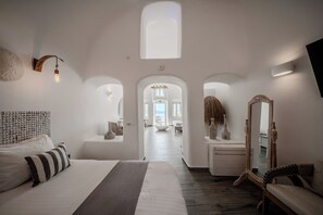 A hotel room with a large bed, a mirror, a wall-mounted light fixture, and an arched doorway leading to a balcony. - Angel Luxury Suites (Santorini)