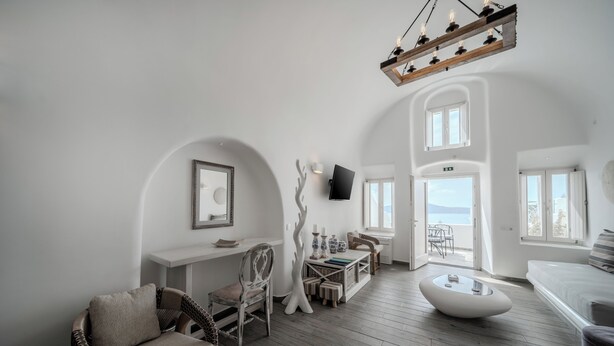 A spacious room with a high ceiling, a wooden floor, a white sofa, a chair, a small table, and a TV mounted on the wall. - Angel Luxury Suites (Santorini)