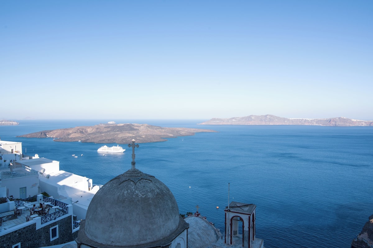 A coastal view with a prominent dome structure, a small bell tower, and a clear blue sea.