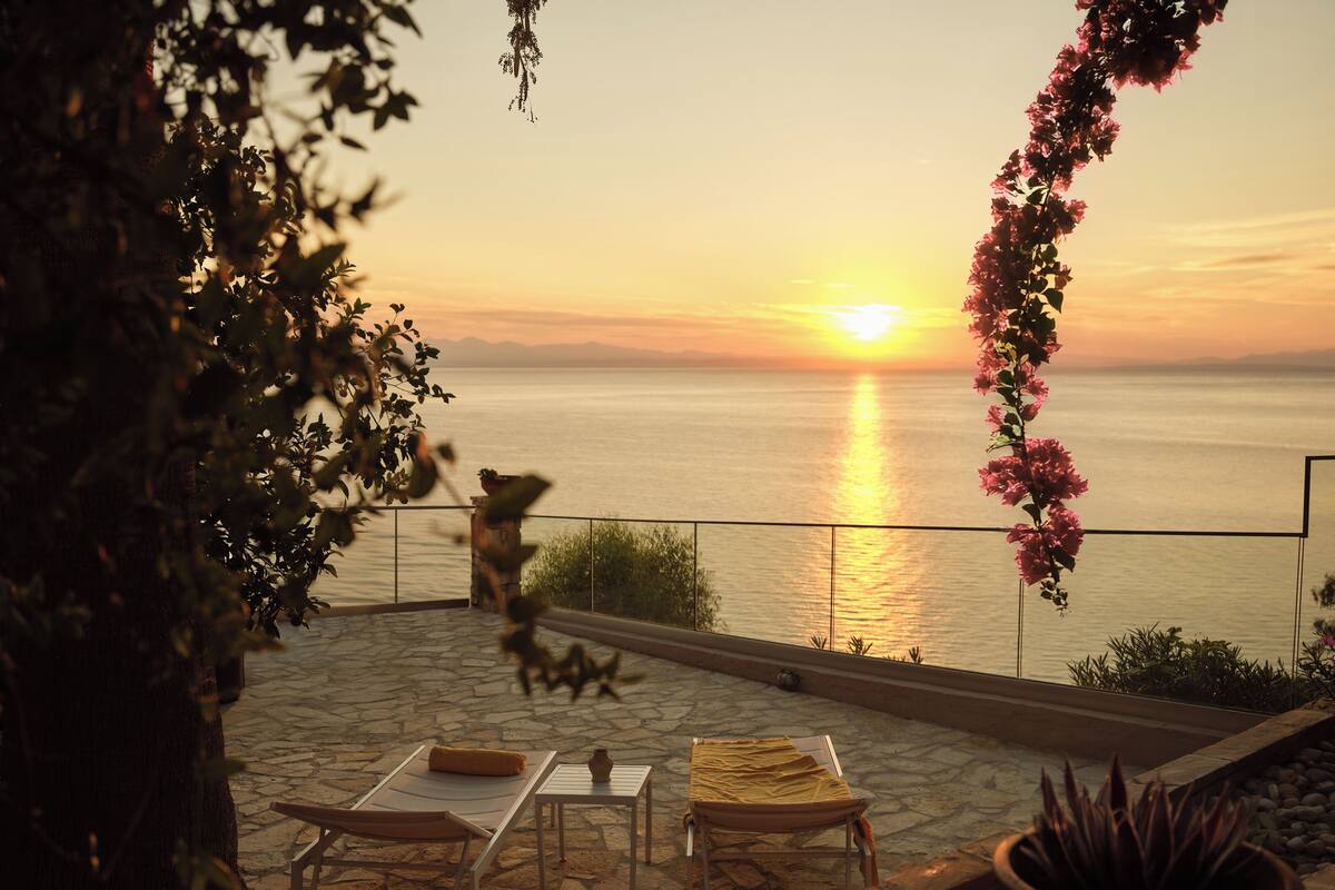 A patio with a view of the sunset over the water, a table with a plant, and a chair.