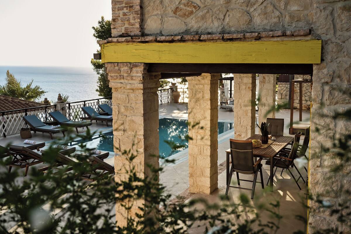 A stone building with a yellow beam, a pool, lounge chairs, and a table with chairs.