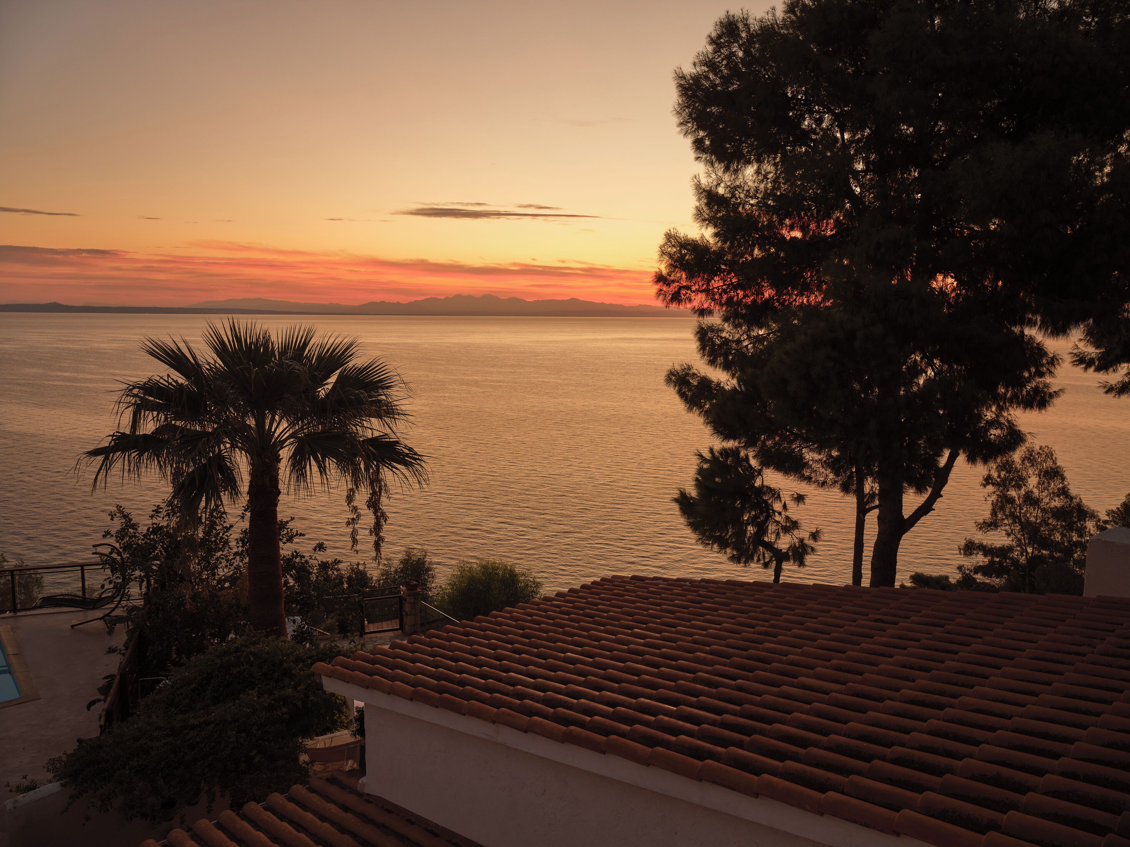 Vista costera con atardecer sobre el agua, palmeras y un tejado de baldosas.