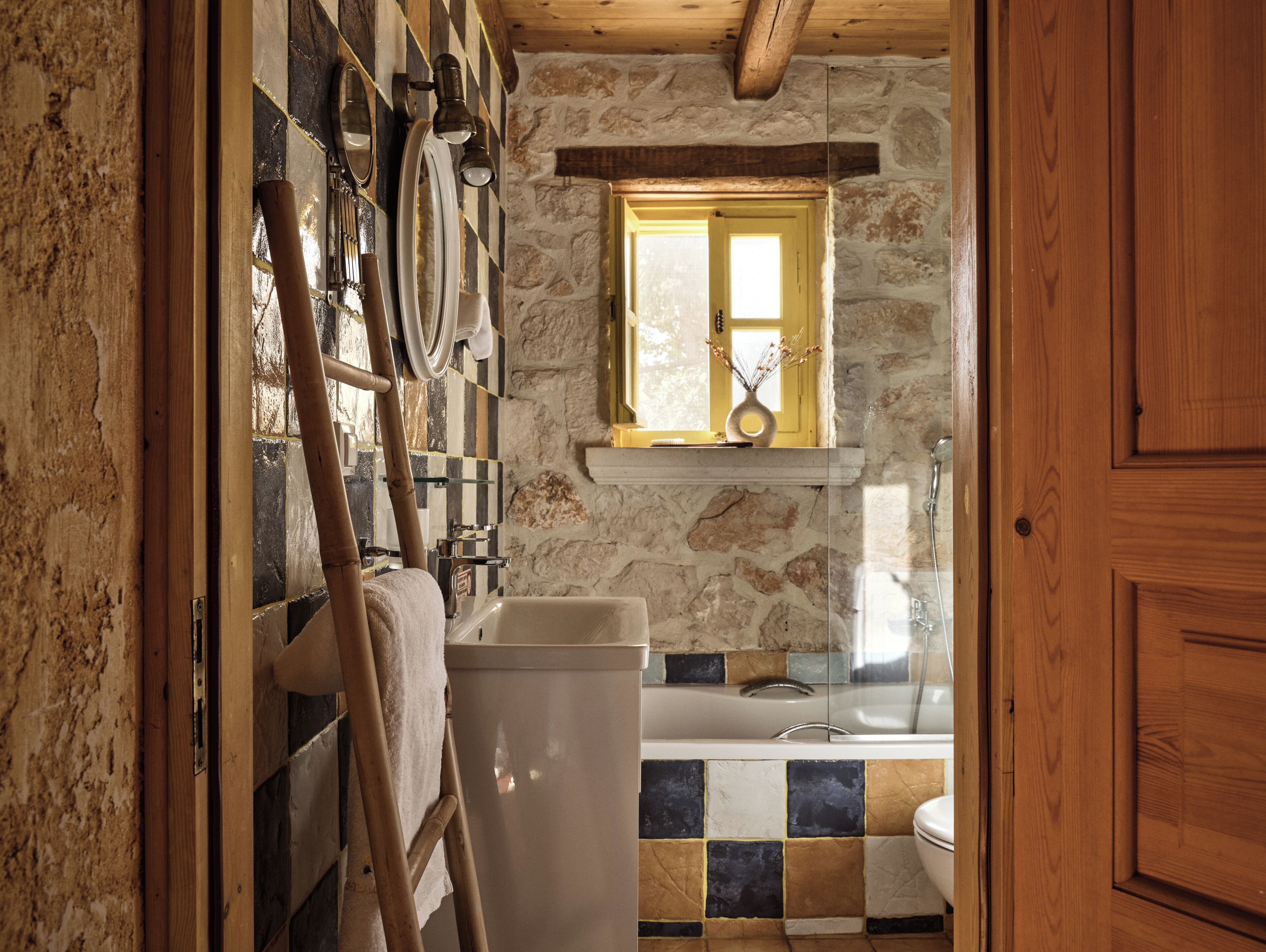 A rustic bathroom with a stone wall, a wooden ladder, a bathtub, and a window with a view.