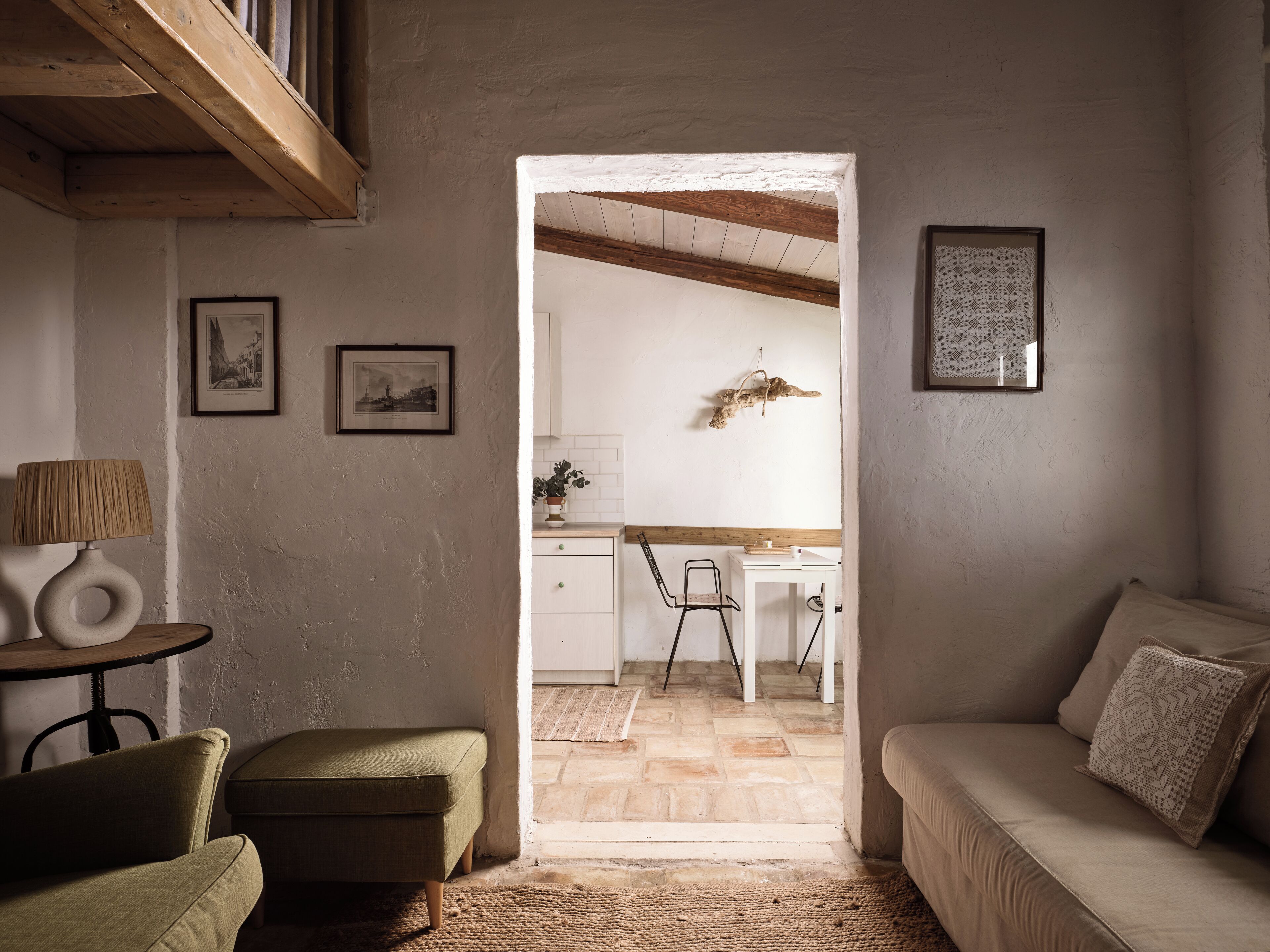 Una sala de estar acogedora con un sillón verde, un sofá beige y una mesita de noche con una lámpara. Un pasillo da a una cocina con techo de madera y un adorno colgante.